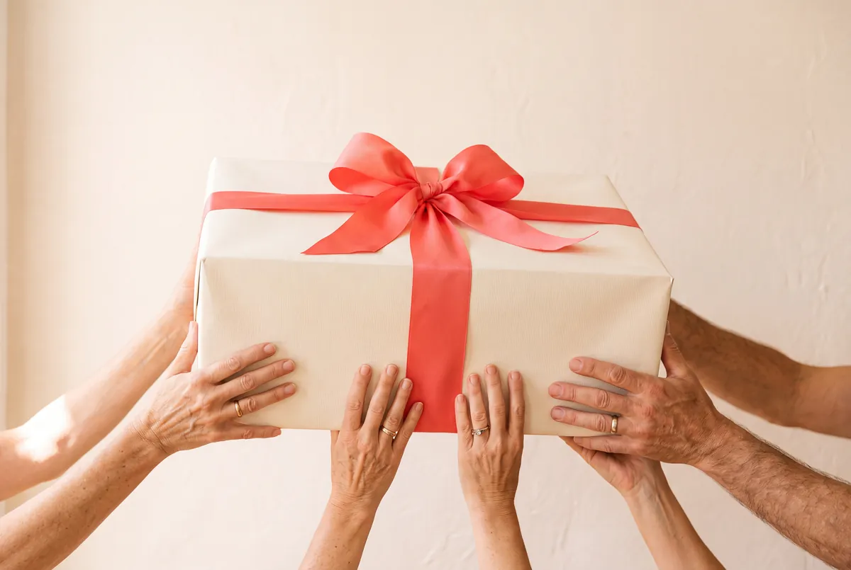 Six hands together holding one large gift box with a wide coral ribbon bow.
